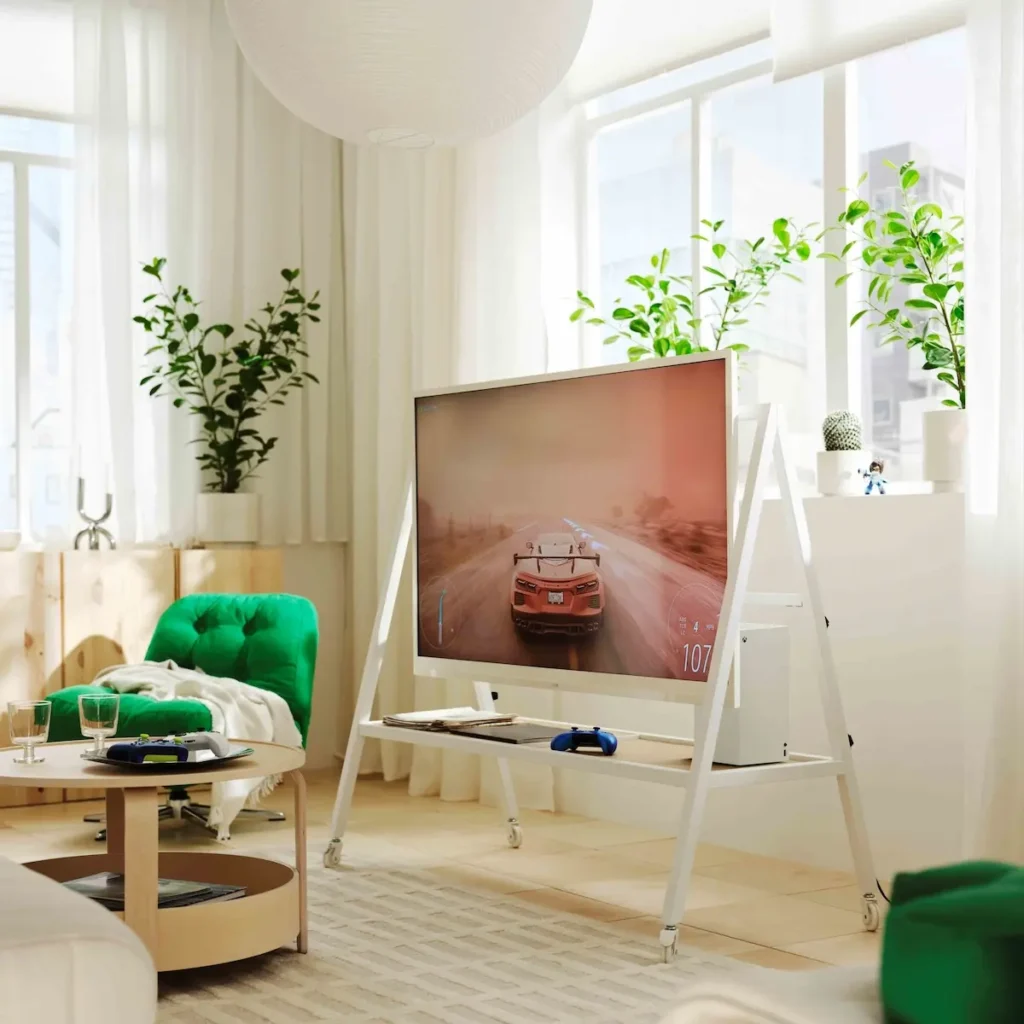 จอเกมบนขาตั้งสีขาวมีล้อในห้องนั่งเล่นที่ตกแต่งด้วยต้นไม้และเก้าอี้สีเขียว